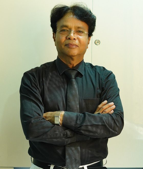A professionally dressed man in a black shirt and tie standing with arms crossed, posing confidently against a light background.