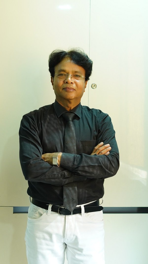 Dr. K. L. Prajapati stands confidently with arms crossed, dressed in a black formal shirt and tie.