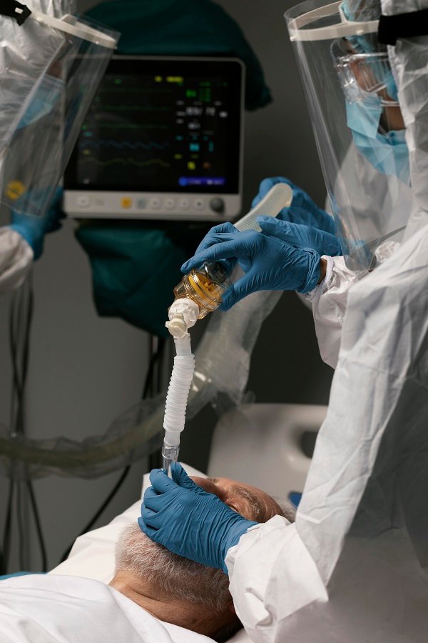 Medical team in protective gear providing respiratory support to a patient lying in a hospital bed with monitoring equipment in the background.